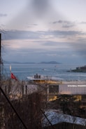 A contemporary building labeled 'Istanbul Modern' sits by a body of water under a cloudy sky. Several boats are visible on the water, and distant islands are seen in the background. A Turkish flag is prominently displayed on a pole near the building. Bare trees are partially framing the view.