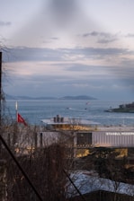 A contemporary building labeled 'Istanbul Modern' sits by a body of water under a cloudy sky. Several boats are visible on the water, and distant islands are seen in the background. A Turkish flag is prominently displayed on a pole near the building. Bare trees are partially framing the view.
