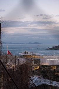 A contemporary building labeled 'Istanbul Modern' sits by a body of water under a cloudy sky. Several boats are visible on the water, and distant islands are seen in the background. A Turkish flag is prominently displayed on a pole near the building. Bare trees are partially framing the view.