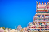 A brightly colored Hindu temple with intricate carvings and ornate details stands prominently against a clear blue sky. The temple exhibits a vibrant array of colors including pinks, blues, greens, and yellows. In the background, additional temple structures and palm trees are visible, creating a picturesque scene.