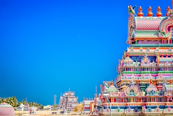 A brightly colored Hindu temple with intricate carvings and ornate details stands prominently against a clear blue sky. The temple exhibits a vibrant array of colors including pinks, blues, greens, and yellows. In the background, additional temple structures and palm trees are visible, creating a picturesque scene.