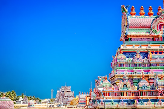 A brightly colored Hindu temple with intricate carvings and ornate details stands prominently against a clear blue sky. The temple exhibits a vibrant array of colors including pinks, blues, greens, and yellows. In the background, additional temple structures and palm trees are visible, creating a picturesque scene.