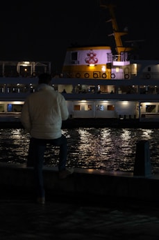 A person wearing a light-colored jacket stands near the edge of a waterfront at night, looking out at a ship with bright lights. The ship has a prominent logo on it and is reflected in the dark water, creating a serene and contemplative scene.