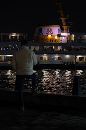 A person wearing a light-colored jacket stands near the edge of a waterfront at night, looking out at a ship with bright lights. The ship has a prominent logo on it and is reflected in the dark water, creating a serene and contemplative scene.