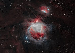 A vibrant and detailed image of a nebula in space, featuring swirling clouds of interstellar dust and gas. The nebula exhibits brilliant shades of red, white, and blue colors with bright stars scattered throughout the dark background. The contrast between the luminous nebula and the surrounding void gives a sense of depth and vastness.
