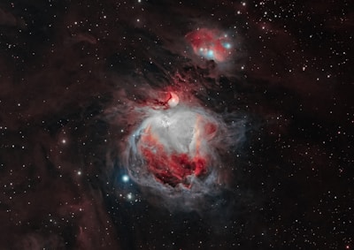 A vibrant and detailed image of a nebula in space, featuring swirling clouds of interstellar dust and gas. The nebula exhibits brilliant shades of red, white, and blue colors with bright stars scattered throughout the dark background. The contrast between the luminous nebula and the surrounding void gives a sense of depth and vastness.