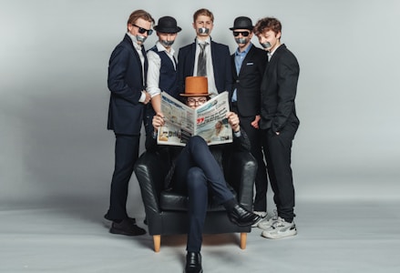 Five individuals dressed in formal suits are positioned in a studio setting with a neutral gray backdrop. Four are standing with tape over their mouths, wearing hats and sunglasses, displaying an air of mystery or secrecy. The fifth person, seated in a black armchair, is reading a newspaper and wearing a brown hat, appearing relaxed and engrossed in the paper.