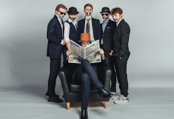 Five individuals dressed in formal suits are positioned in a studio setting with a neutral gray backdrop. Four are standing with tape over their mouths, wearing hats and sunglasses, displaying an air of mystery or secrecy. The fifth person, seated in a black armchair, is reading a newspaper and wearing a brown hat, appearing relaxed and engrossed in the paper.