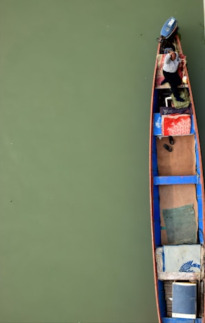 A narrow boat on green water with a person sitting at the end, featuring a small motor and various items arranged neatly on the deck, such as a pair of shoes and colorful cushions.