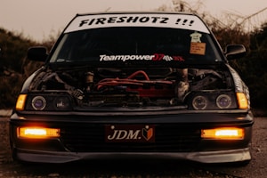 A car with its hood removed, exposing its engine. The windshield displays various stickers, including 'FIRESHOTZ !!!' at the top and 'Teampower07' underneath. The headlights are on, casting a soft glow in a dimly lit environment. The front license plate reads 'JDM'. Surrounding vegetation is visible in the background.