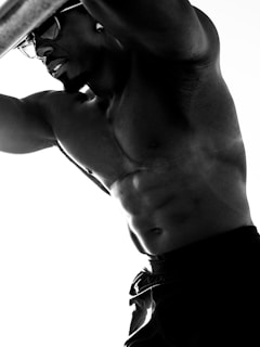 A black and white photograph depicts a muscular man with an athletic build. He is shirtless, wearing dark shorts or pants, and is positioned in a pose that highlights his toned physique. The lighting accentuates the contours of his muscles, creating strong contrasts and shadows. He appears to be wearing glasses and looking contemplatively into the distance.
