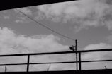 Black and white photo featuring a cloudy sky with a metal railing and a security camera mounted on a pole in the foreground. A wire runs diagonally across the image.