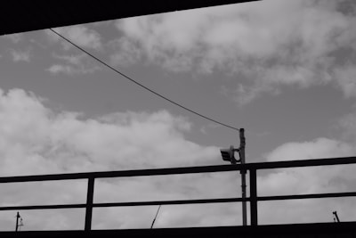 Black and white photo featuring a cloudy sky with a metal railing and a security camera mounted on a pole in the foreground. A wire runs diagonally across the image.