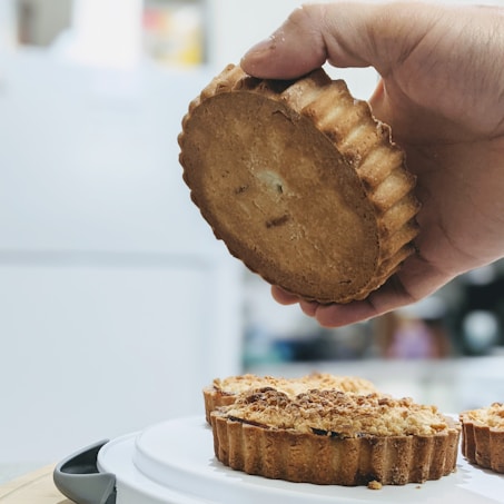 A hand holding a round, fluted-edged pastry or tart with a golden-brown crust. Below are additional pastries resting on a white plate or tray. The pastries have a crumbly texture on top.