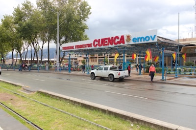 A bus terminal entrance with a sign displaying 'Terminal Terrestre Cuenca' and 'emov'. Several people are walking near the entrance, and a white pickup truck is moving on the road. Trees line the background, and there are flags hanging above the entrance.