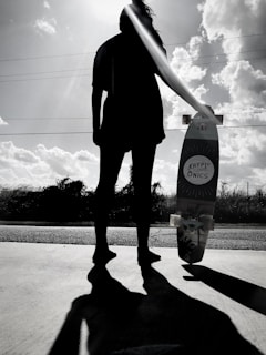 A silhouette of a person standing on a paved surface holding a skateboard with the brand name 'Kryptonics' visible. The sun is positioned behind the individual's head, creating a dramatic backlighting effect. Fluffy clouds are scattered across the bright sky, and low vegetation is visible in the background.