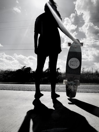 A silhouette of a person standing on a paved surface holding a skateboard with the brand name 'Kryptonics' visible. The sun is positioned behind the individual's head, creating a dramatic backlighting effect. Fluffy clouds are scattered across the bright sky, and low vegetation is visible in the background.