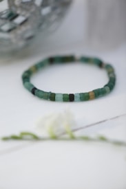 A beaded bracelet made up of small, cube-shaped beads in various shades of green and a touch of tan sits on a light-colored surface. The background features a decorative object with a reflective texture, and there is a blurred floral element in the foreground.