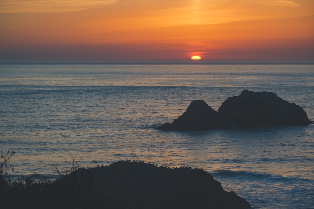 the sun is setting over the ocean with rocks in the foreground