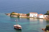 A serene coastal scene with a wooden boat sailing near a small island. The island features charming white Mediterranean-style buildings with tile roofs. Clear turquoise water surrounds the island, reflecting the vibrant colors of the landscape. Lush greenery is present on the island, enhancing the picturesque view.
