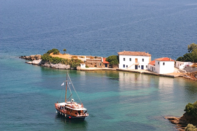A serene coastal scene with a wooden boat sailing near a small island. The island features charming white Mediterranean-style buildings with tile roofs. Clear turquoise water surrounds the island, reflecting the vibrant colors of the landscape. Lush greenery is present on the island, enhancing the picturesque view.