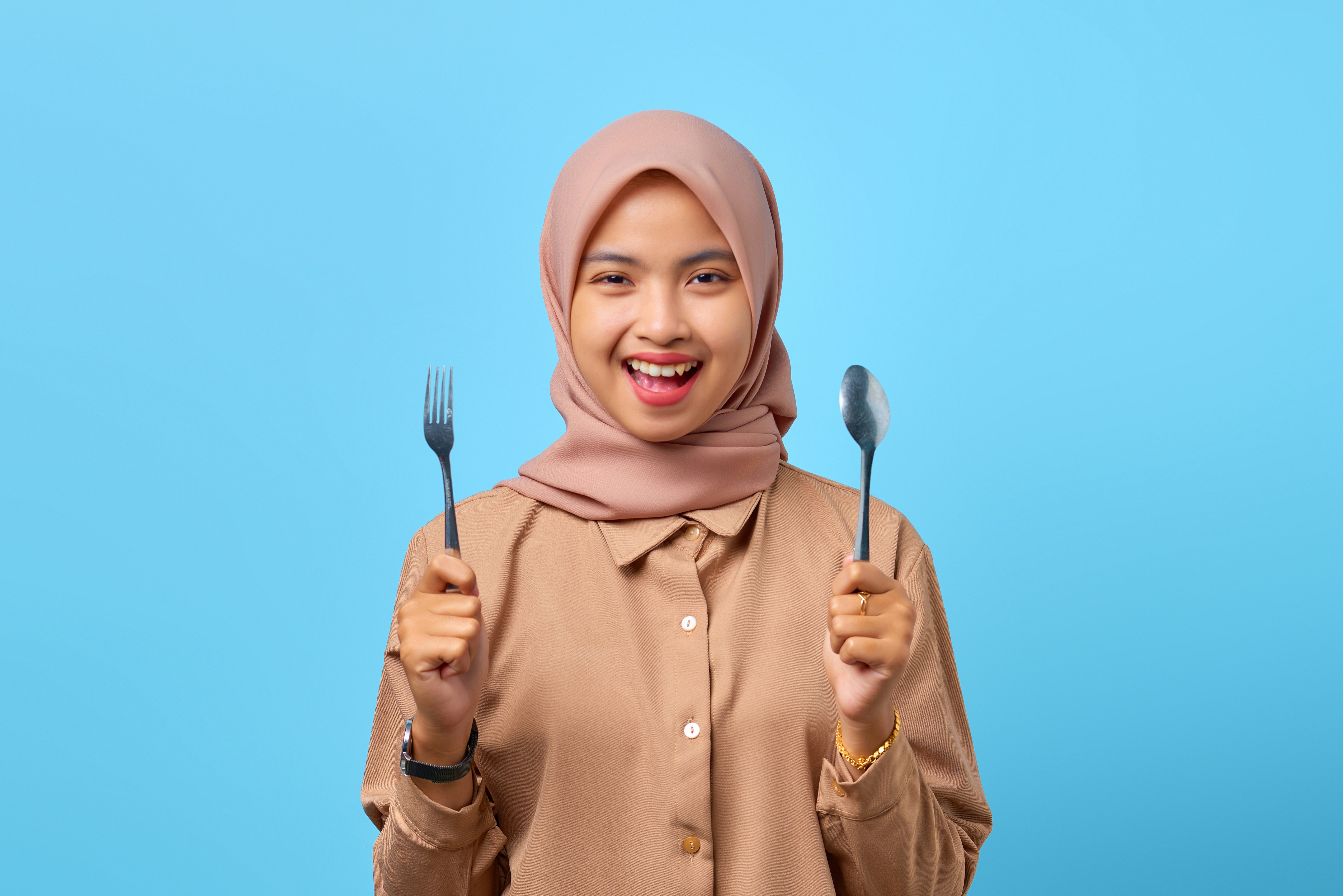 a woman in a hijab holding two forks and a knife, Portrait of smiling young Asian woman hold fork and spoon over blue background
