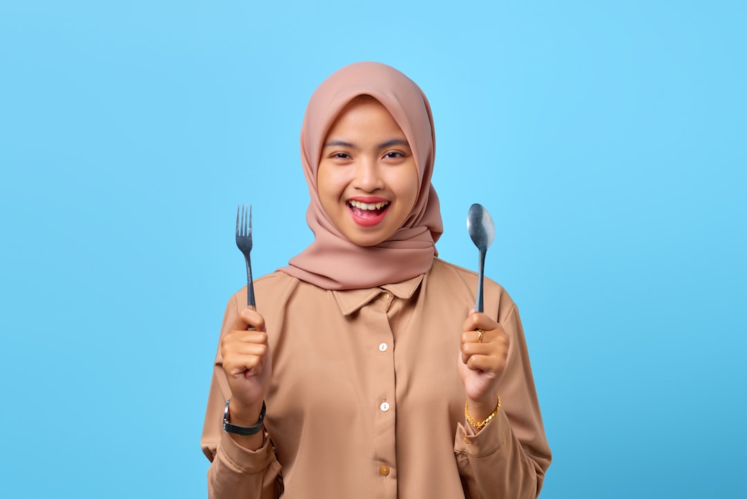 a woman in a hijab holding two forks and a knife, Portrait of smiling young Asian woman hold fork and spoon over blue ba
