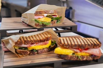 Three sandwiches are arranged on a wooden stand, each filled with ingredients like ham, scrambled eggs, lettuce, and possibly cheese, presented in brown paper wrapping. The sandwiches appear to be made with hearty, whole grain bread, and are located in a kitchen setting.