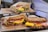 Three sandwiches are arranged on a wooden stand, each filled with ingredients like ham, scrambled eggs, lettuce, and possibly cheese, presented in brown paper wrapping. The sandwiches appear to be made with hearty, whole grain bread, and are located in a kitchen setting.