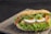 A sandwich with a seeded, crusty bread roll filled with layers of fresh lettuce, slices of boiled eggs, red bell peppers, crispy chicken, and a drizzle of creamy sauce. The ingredients are vibrant and arranged neatly, suggesting a rich, flavorful meal.