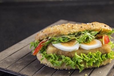 A sandwich with a seeded, crusty bread roll filled with layers of fresh lettuce, slices of boiled eggs, red bell peppers, crispy chicken, and a drizzle of creamy sauce. The ingredients are vibrant and arranged neatly, suggesting a rich, flavorful meal.