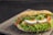 A sandwich with a seeded, crusty bread roll filled with layers of fresh lettuce, slices of boiled eggs, red bell peppers, crispy chicken, and a drizzle of creamy sauce. The ingredients are vibrant and arranged neatly, suggesting a rich, flavorful meal.