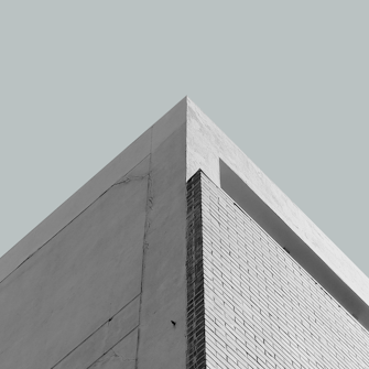 A minimalist architectural photograph focusing on the sharp corner of a building, with clean lines and angles. The facade consists of a combination of smooth plaster and brick textures, set against a muted light blue sky, creating a sense of simplicity and modernity.