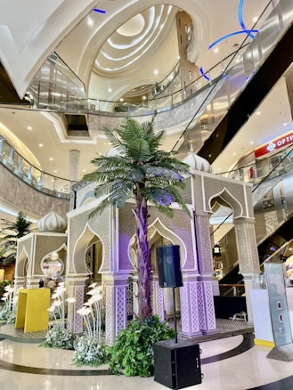 A beautifully decorated shopping mall interior featuring a large ornate structure with intricate patterns, possibly representing an architectural theme. The setting includes palm trees, elaborate lighting, and multiple levels of glass railings and escalators. Ambient and architectural lighting complement the sophisticated design.