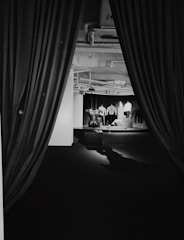 A monochrome view of an interior space framed by large curtains. Inside, mannequins display various garments against a dark backdrop, with one visible wall and ceiling pipes overhead.