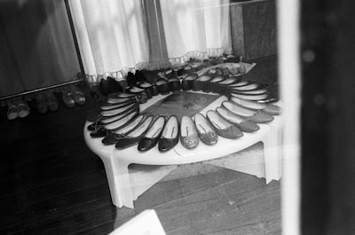 A display of various ballet flats is arranged in a semi-circle on a small table inside a store. The shoes feature different styles and textures, including glossy and sparkly finishes. The background includes additional shoes lined up against the wall, and curtains are drawn behind the arrangement.