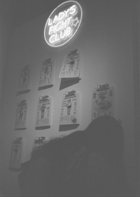 A monochrome image features a wall adorned with a neon sign reading 'Lady's Fight Club.' Beneath the sign, a series of vintage-style posters is clipped to the wall, each featuring illustrations and text. The scene is captured in low light, adding a moody and nostalgic atmosphere.