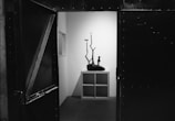 A minimalistic gallery space is visible through a partially open door. Inside, a sculptural piece featuring an abstract figure standing on a textured base is placed on a white cubic shelf. The sculpture casts a distinct, elongated shadow on the plain white wall behind it.