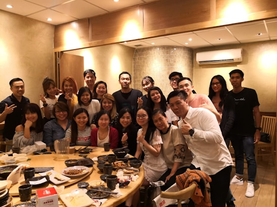 A warm, candid photo of six men sharing a meal and laughter around a community center table.