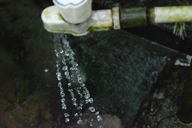 Water droplets spill from a pipe or tap, against a dark, mossy background. The droplets are captured in motion, showcasing their clarity and the dynamics of flowing water.
