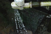Water droplets spill from a pipe or tap, against a dark, mossy background. The droplets are captured in motion, showcasing their clarity and the dynamics of flowing water.