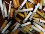 A pile of used cigarette butts and ashes submerged in a liquid, with a variety of brands visible. The butts are wet, and some are partially burnt. The liquid has a brownish hue, and there are small debris and particles scattered around.