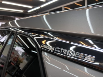 A detailed view of a car badge featuring the text 'Corolla Cross' on the side of a vehicle. The background shows part of an indoor setting with modern lighting and blurred objects.