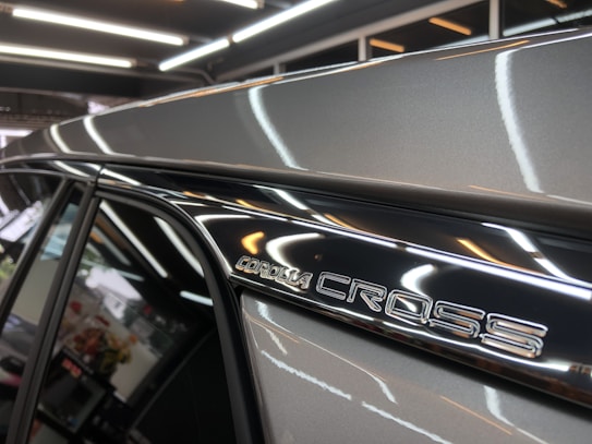 A detailed view of a car badge featuring the text 'Corolla Cross' on the side of a vehicle. The background shows part of an indoor setting with modern lighting and blurred objects.