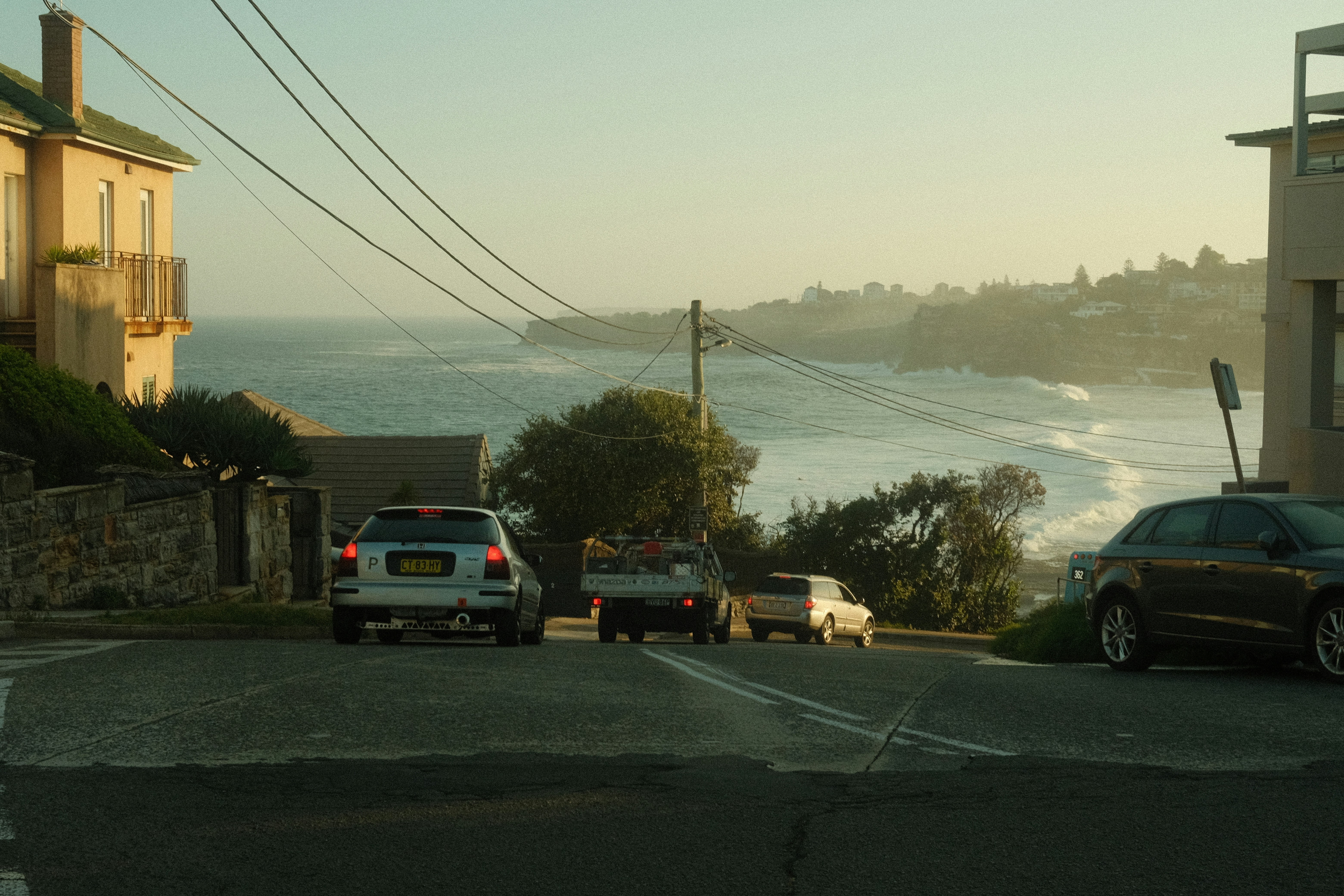 Coastal street scene with parked cars leading toward a sunlit ocean horizon at golden hour.