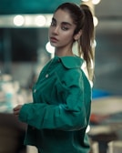 A young woman with light skin, dark eyes, and dark hair pulled back into a ponytail wears a green jacket. She is standing indoors against a blurred background with circular lights, creating a bokeh effect.
