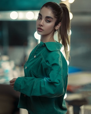 A young woman with light skin, dark eyes, and dark hair pulled back into a ponytail wears a green jacket. She is standing indoors against a blurred background with circular lights, creating a bokeh effect.