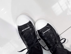 A pair of black and white sneakers with the brand name 'Christian Dior' displayed on the tongue. The shoes have white rubber soles and white toe caps, and they are laced with black shoelaces. They rest on a light-colored tiled floor reflecting some light.