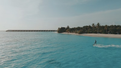 A person surfing on clear, turquoise waters near a tropical beach with lush palm trees. A row of overwater bungalows is visible in the distance, under a bright, clear sky.