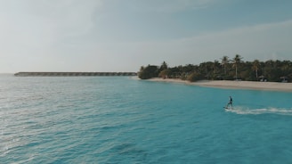 A person surfing on clear, turquoise waters near a tropical beach with lush palm trees. A row of overwater bungalows is visible in the distance, under a bright, clear sky.